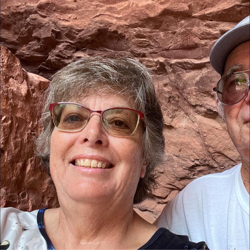 Woman with glasses smiling, next to a man, in front of a red rock formation.