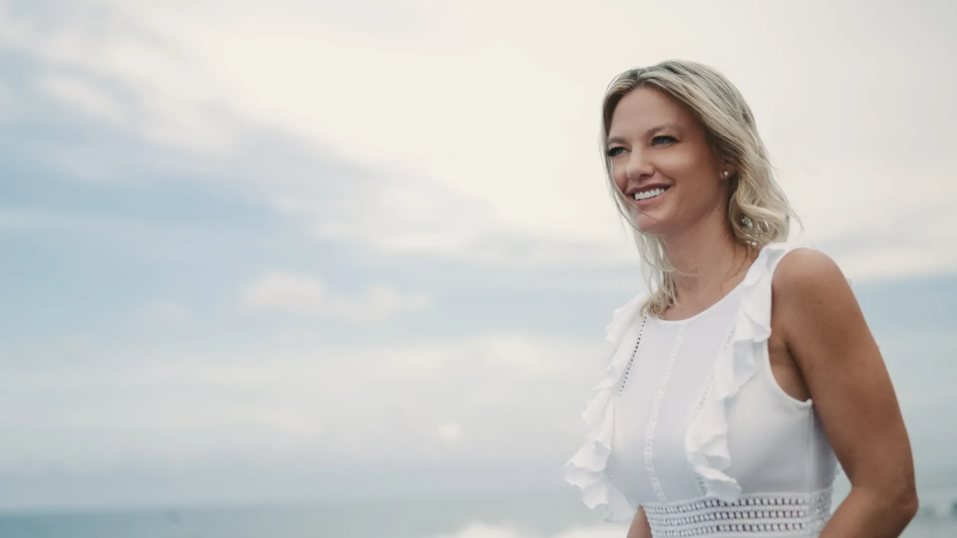 Woman with blonde hair smiles, wearing a white dress, outdoors against a cloudy sky.