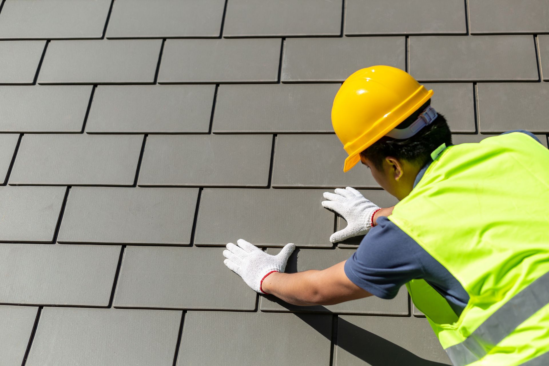 Construction worker installing gray roof shingles wearing safety helmet and vest.