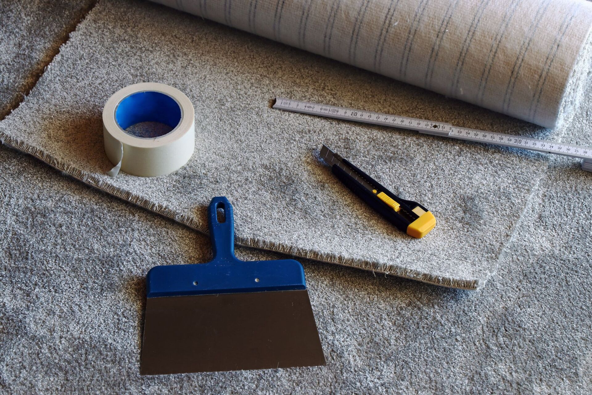 Assorted tools for carpet repair, including a utility knife, carpet adhesive, and a measuring tape.