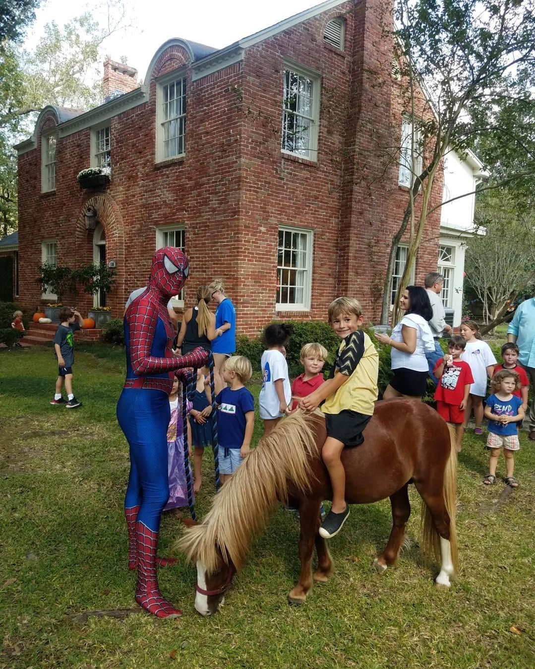 A man dressed as spiderman is talking to a child riding a pony