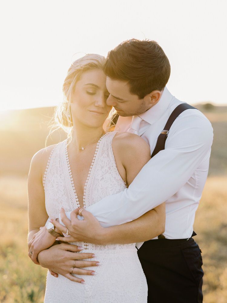 Bride and groom embrace, smiling. Bride in white dress, groom in blue suit, standing outdoors with vineyard background.