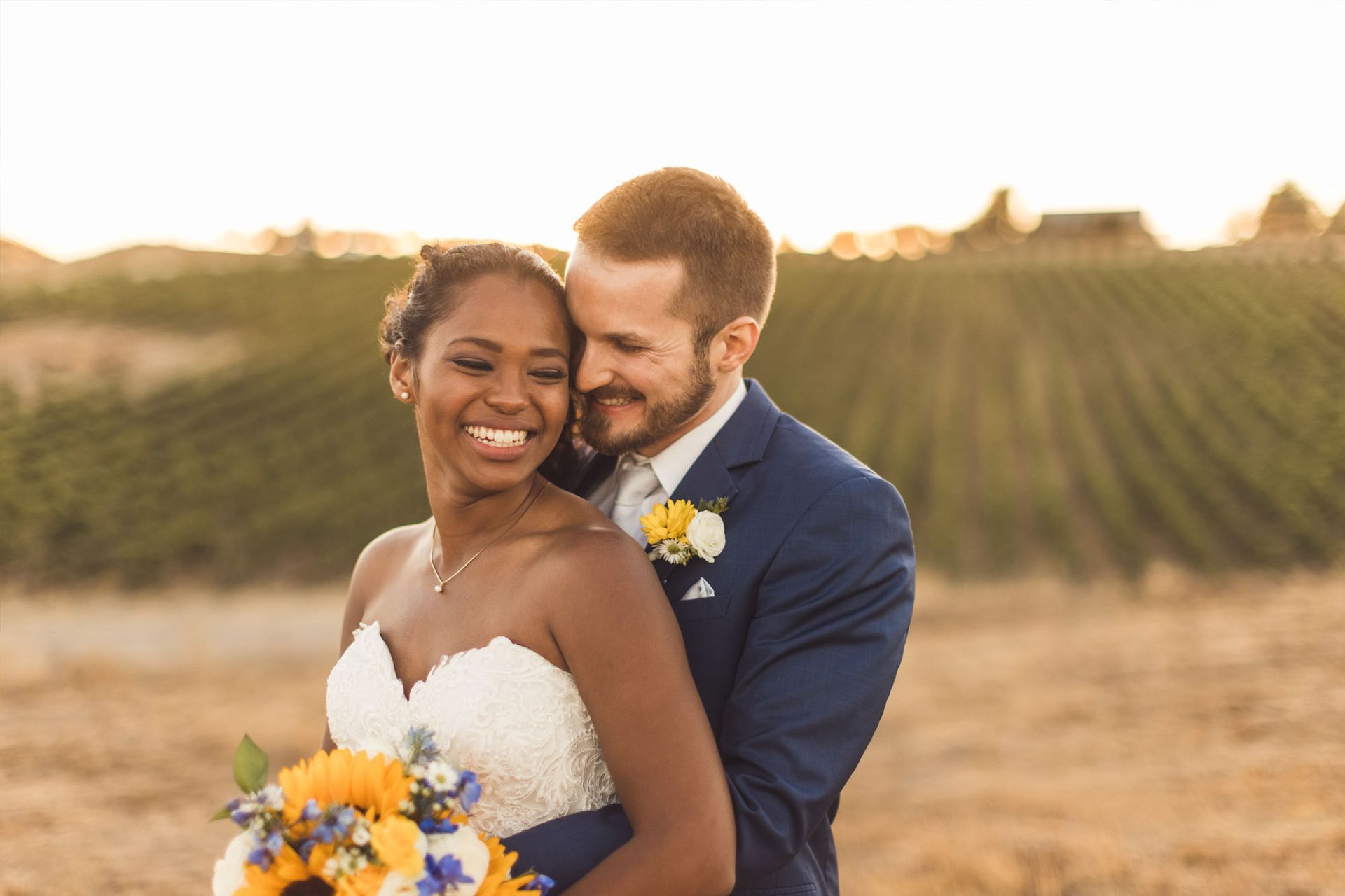 Bride and groom embrace, smiling. Bride in white dress, groom in blue suit, standing outdoors with vineyard background.
