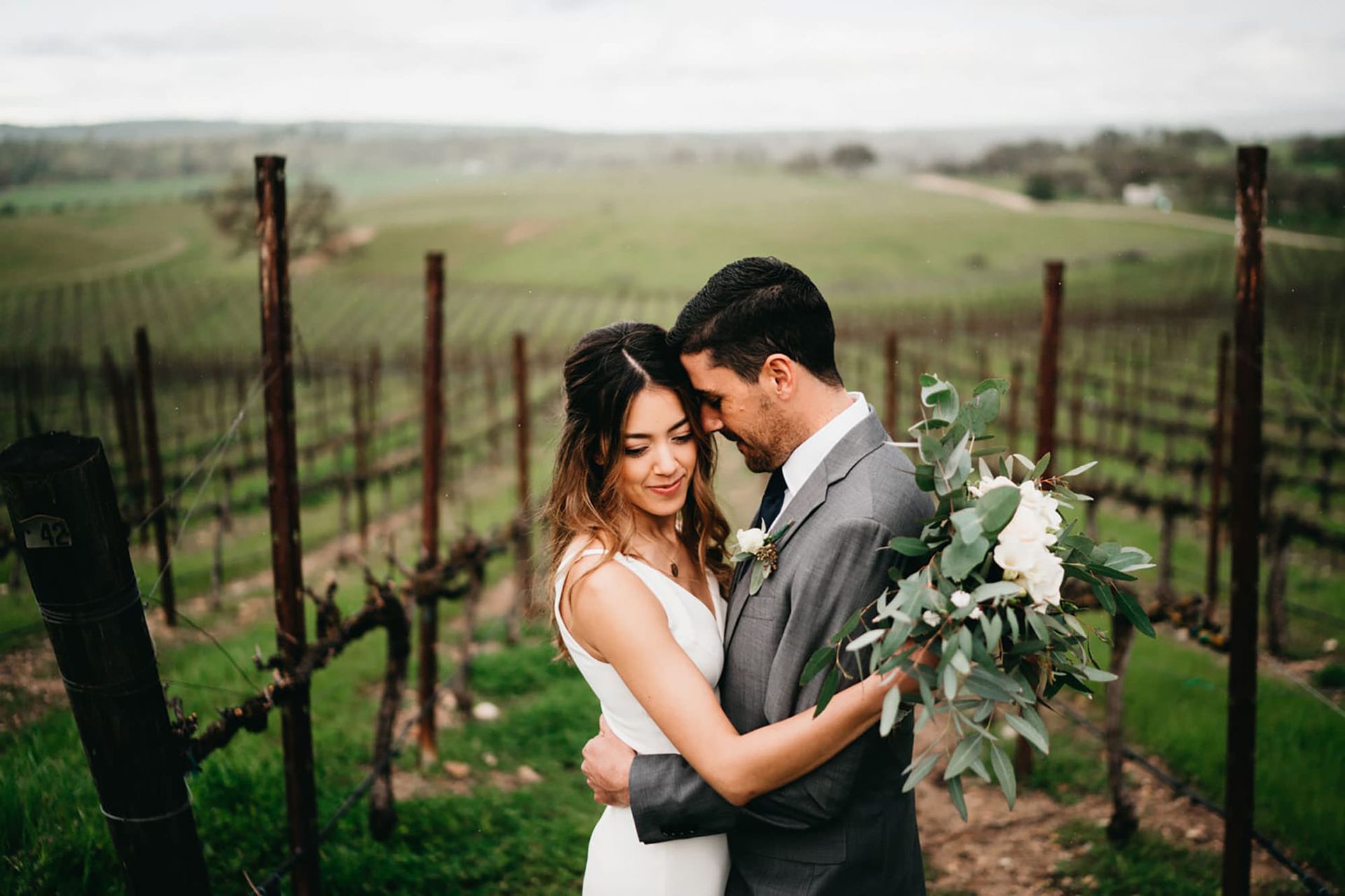 Bride and groom embrace in a vineyard, facing each other. She wears a white dress; he wears a gray suit. Cloudy day.