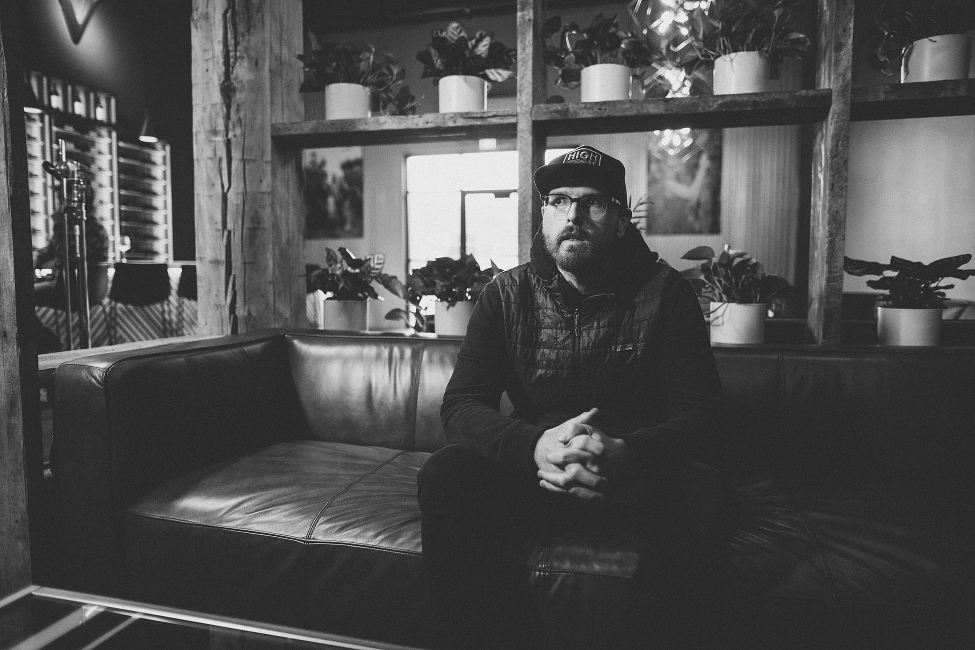 Man in hat and sweater sits on leather sofa, hands clasped, indoors. Plants on shelves in background.
