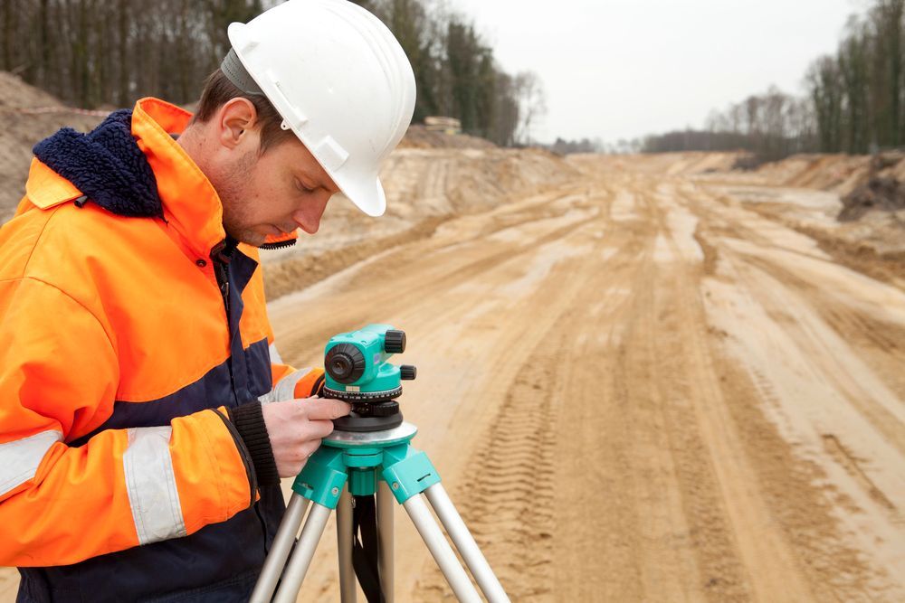 Un trabajador de la construcción con casco y chaqueta de alta visibilidad opera un instrumento de medición en un sitio arenoso.