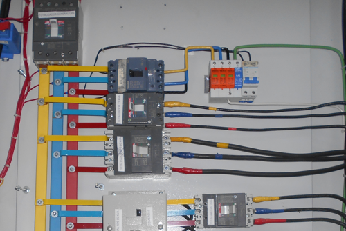Un panel de control eléctrico con varios interruptores, barras colectoras de colores y cableado interno sobre una carcasa blanca.