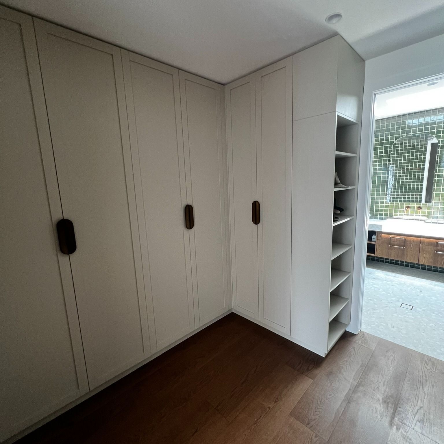 White cabinets with dark handles in a room with wood flooring, adjacent to a bathroom. — Onyx Detailed Joinery in Unanderra, NSW