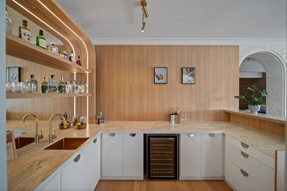 Modern home bar with wood accents, featuring shelves with bottles, glasses, gold faucets, mini fridge — Onyx Detailed Joinery In Unanderra, NSW