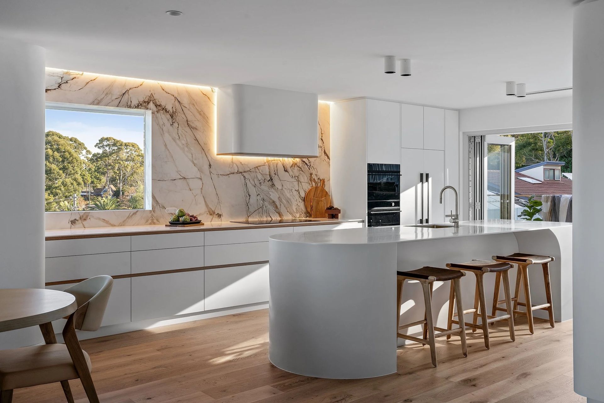 Modern kitchen with sleek white cabinets and a curved island with wooden stools — Onyx Detailed Joinery In Unanderra, NSW