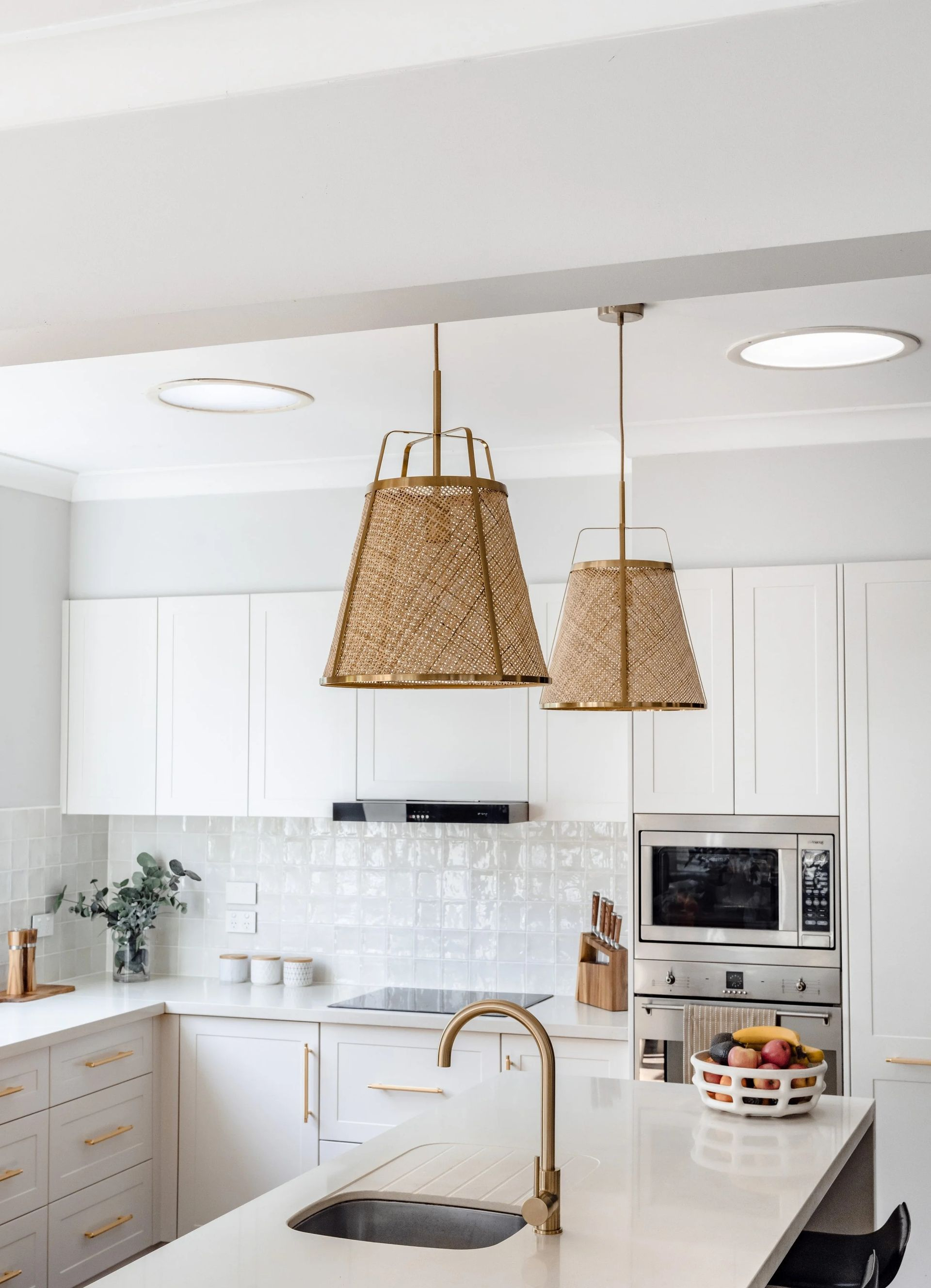 Bright white kitchen with two woven pendant lights over a countertop with a gold faucet. — Onyx Detailed Joinery in Unanderra, NSW