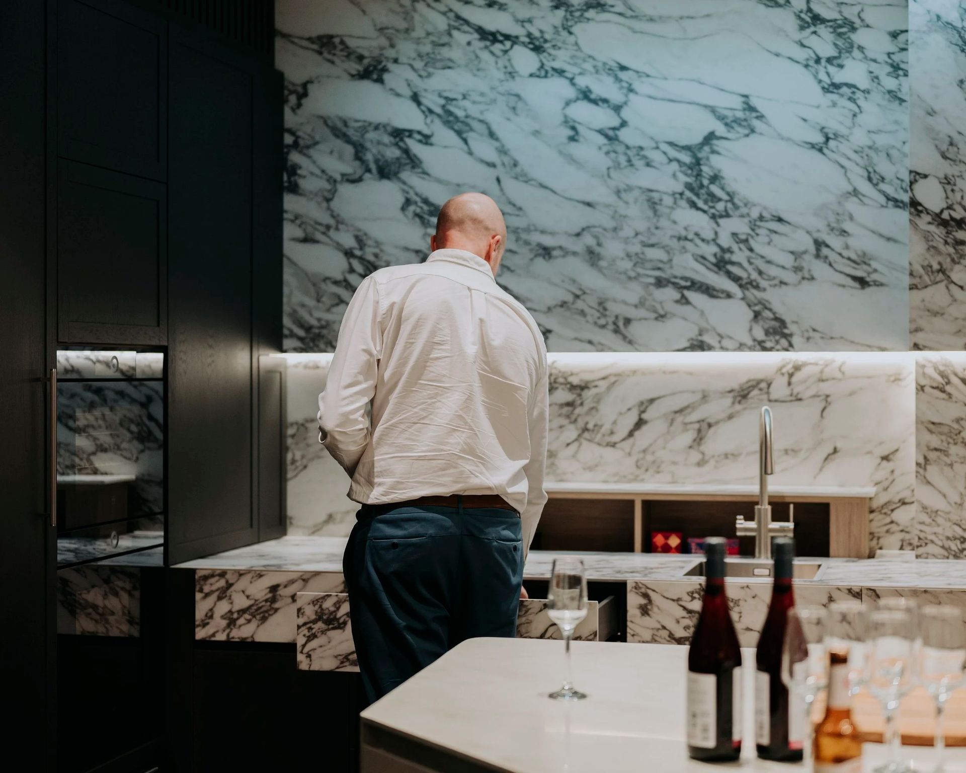 Man standing at a marble countertop in a kitchen; two wine bottles and a glass sit nearby. — Onyx Detailed Joinery in Unanderra, NSW
