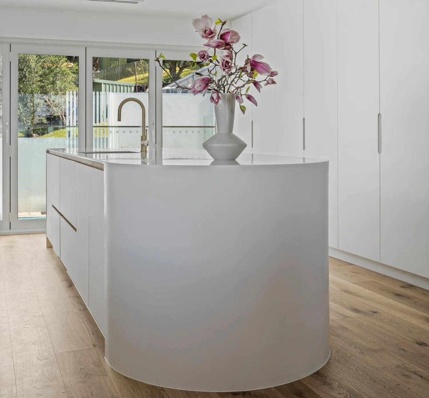 Modern white kitchen with curved island, flowers, and wooden floor.— Onyx Detailed Joinery in Unanderra, NSW