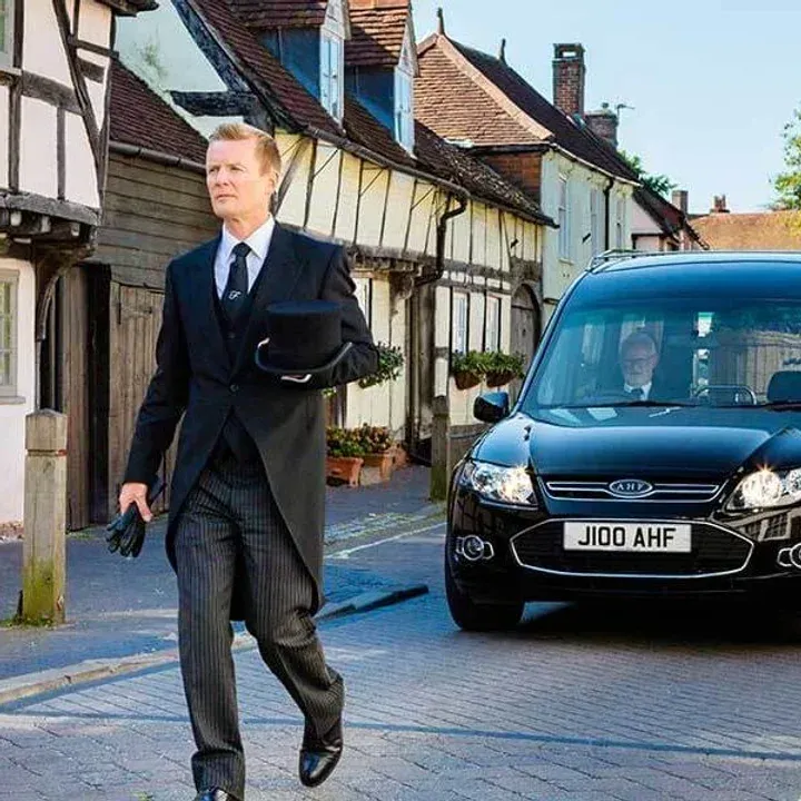 A man in a suit is walking down a street next to a black car with a license plate that says jido ahf