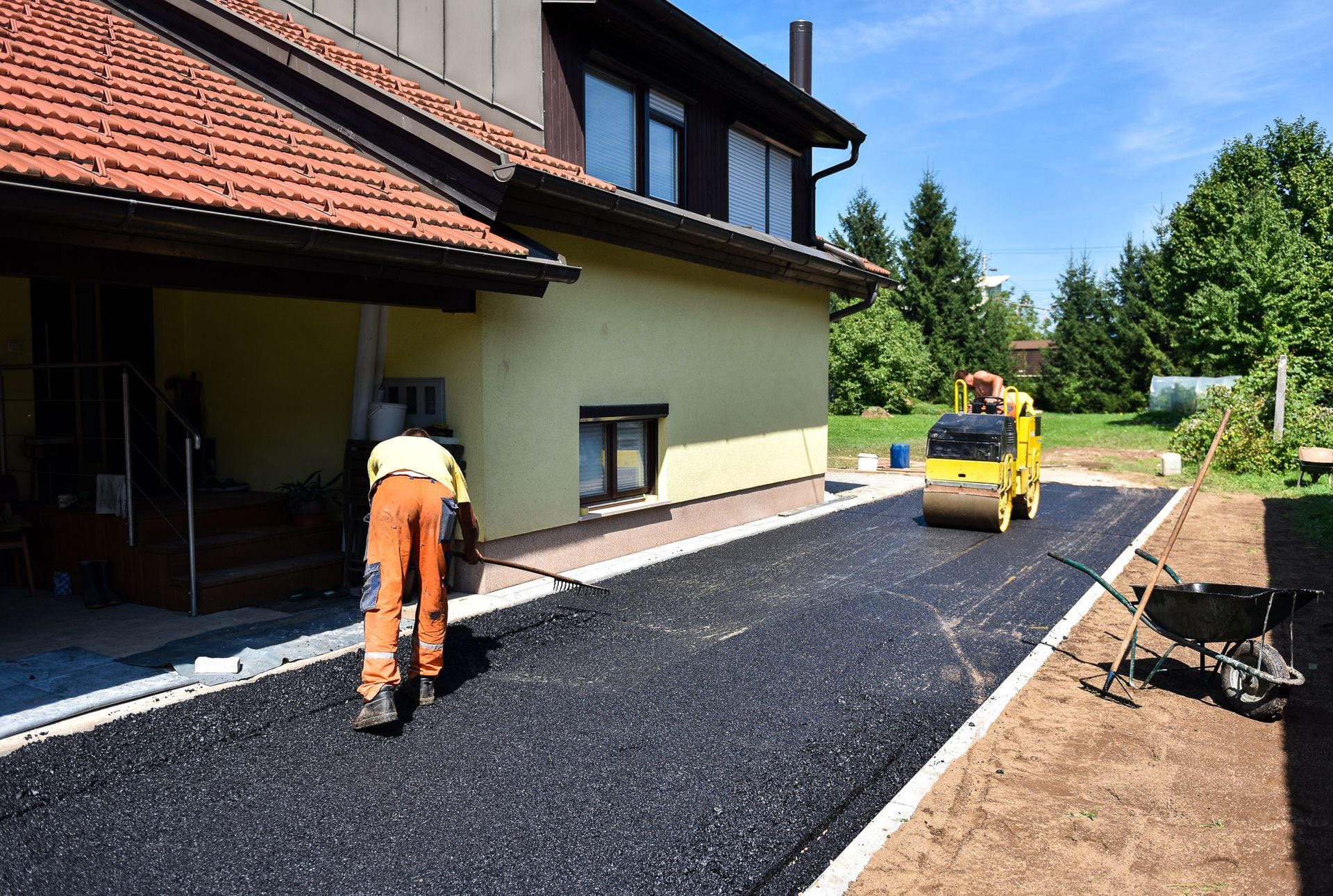 A worker uses a rake to smooth fresh asphalt along the side of a house, while a yellow steamroller waits nearby.