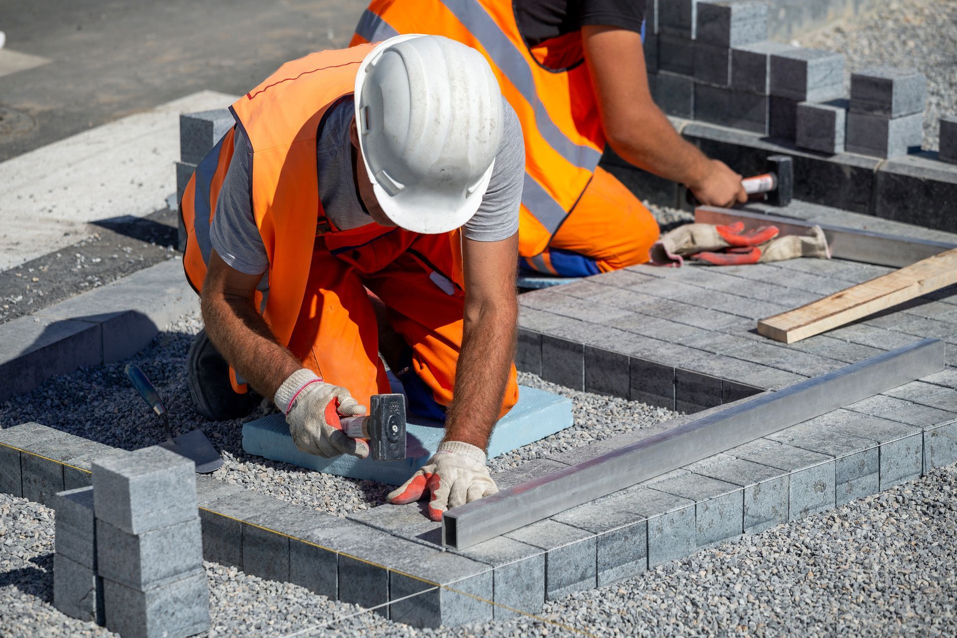 Two workers in high-visibility orange vests and hard hats install stone pavers on a gravel ground.