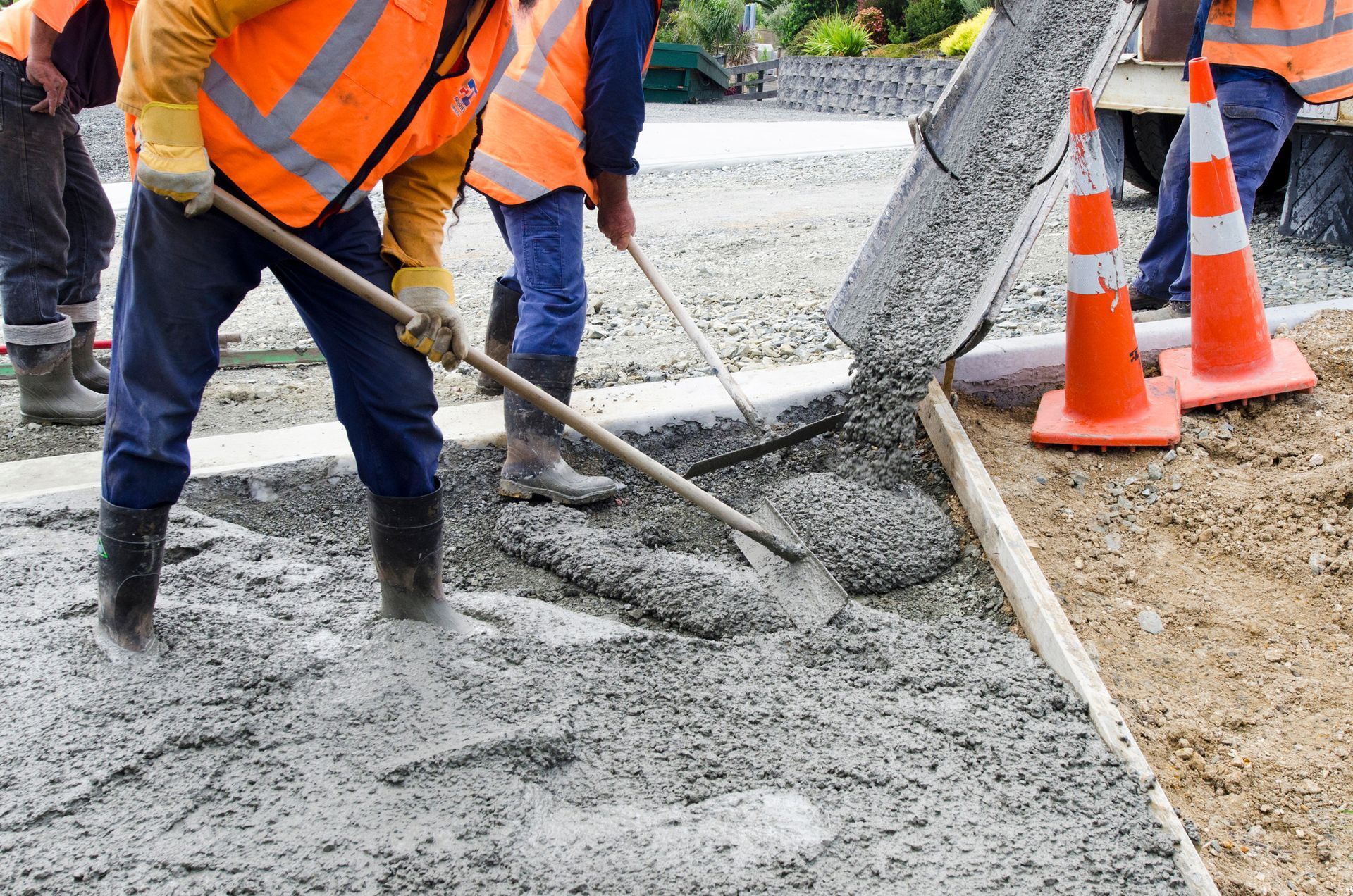 Construction workers in safety vests spread wet concrete from a truck chute at a job site with orange traffic cones.