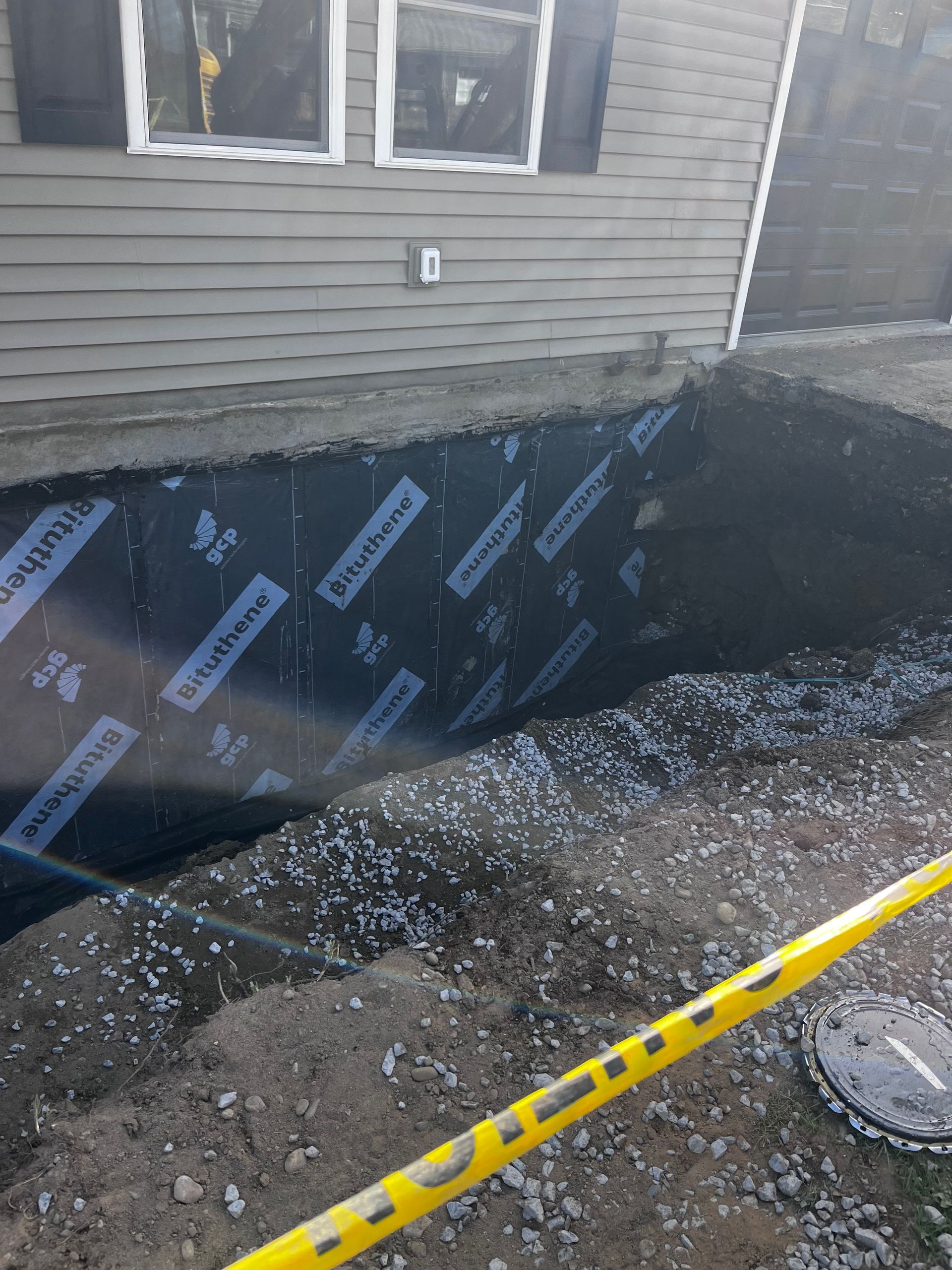 Black waterproofed foundation wall beside a house, with gravel trench and yellow caution tape.