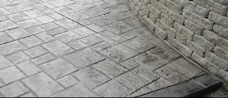 A person wearing work gloves kneels while installing gray paving stones along a curb.