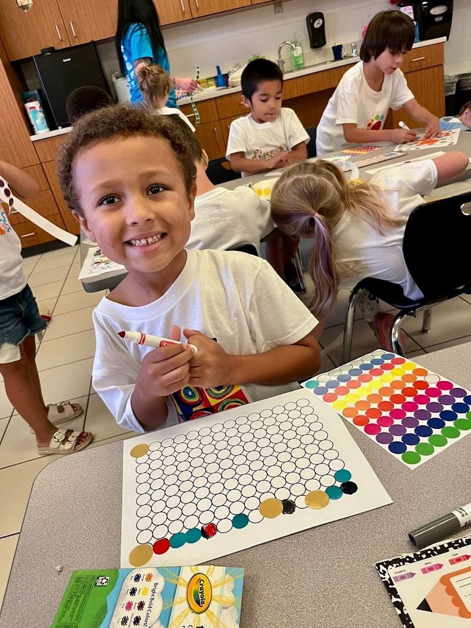 A child smiles while coloring a worksheet at a table in a classroom. Other children work in the background.