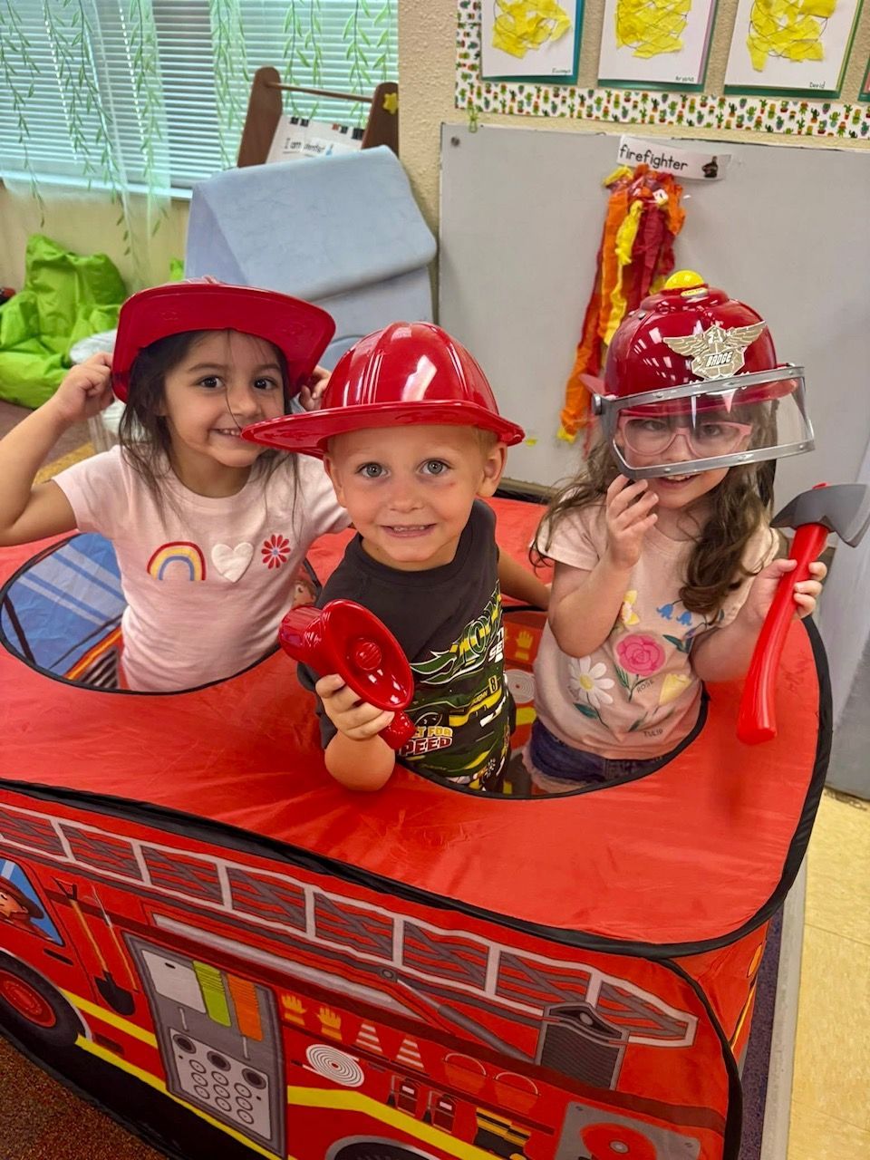 Three children in fire hats play in a fire truck toy, smiling.