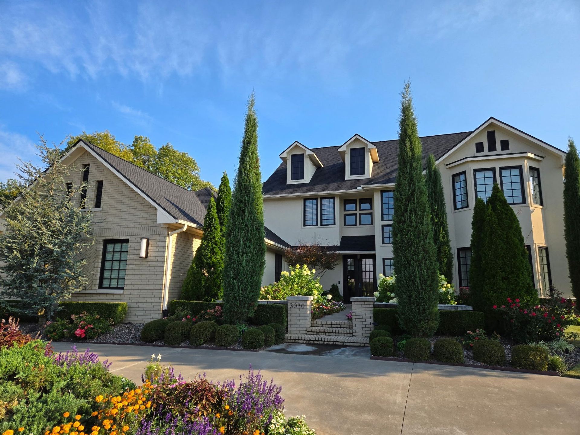 Elegant two-story house with stone facade, manicured front yard, tall evergreen trees, and a blue sky backdrop.