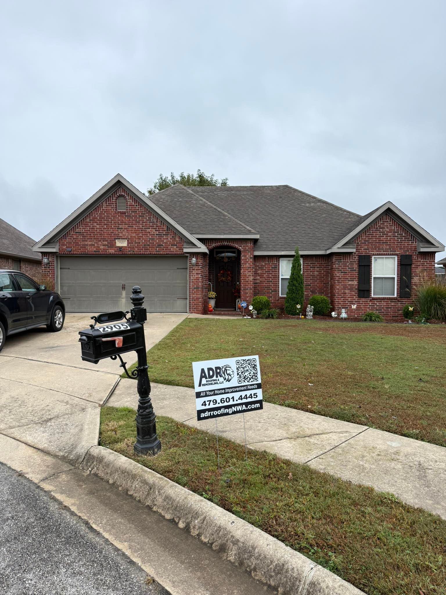 Brick house with dark roof and attached garage. A black mailbox in front, and a sign on the lawn.