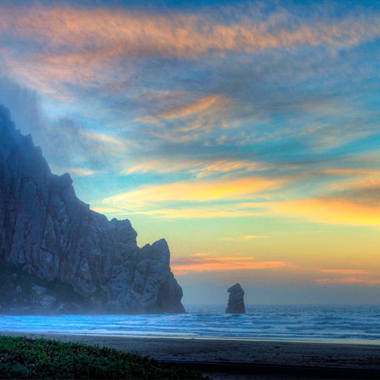A beach with a large rock in the middle of the ocean at sunset.
