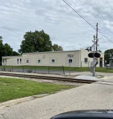 Small beige building beside railroad tracks with a crossing signal and cloudy sky