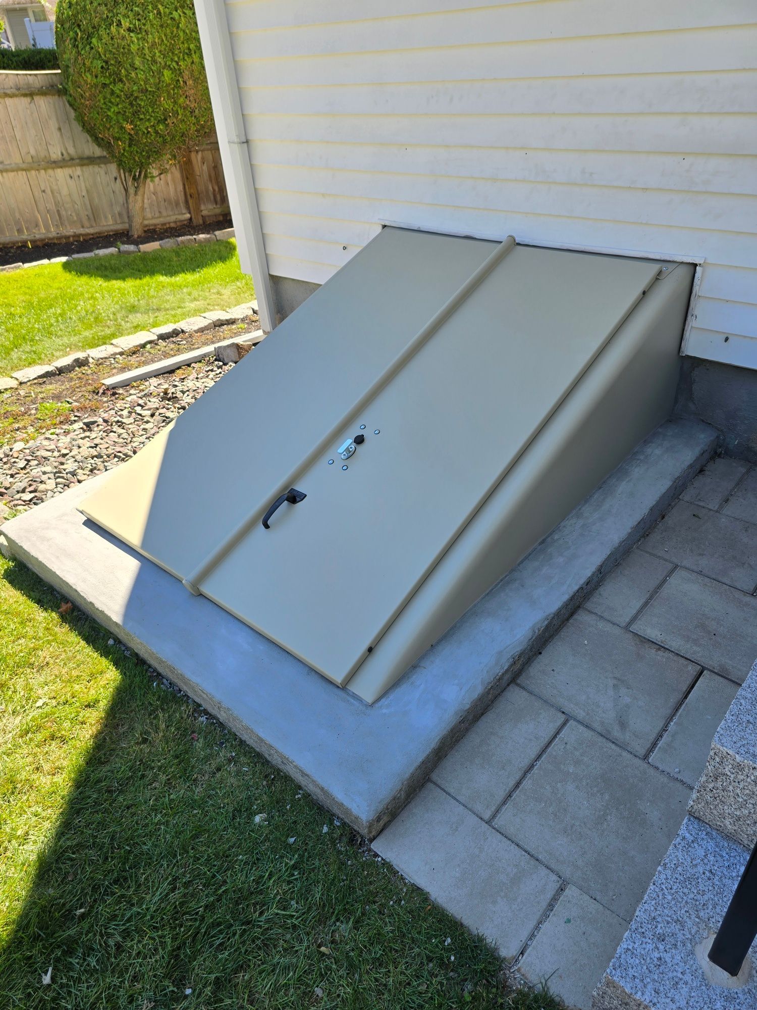 Tan metal basement door on concrete steps, next to a white house.