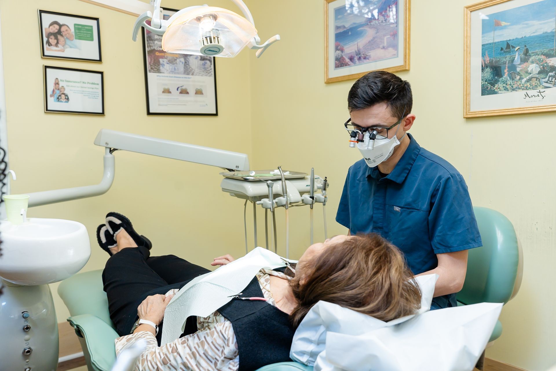 Dentist examining a patient in a dental chair; medical equipment visible, yellow walls.
