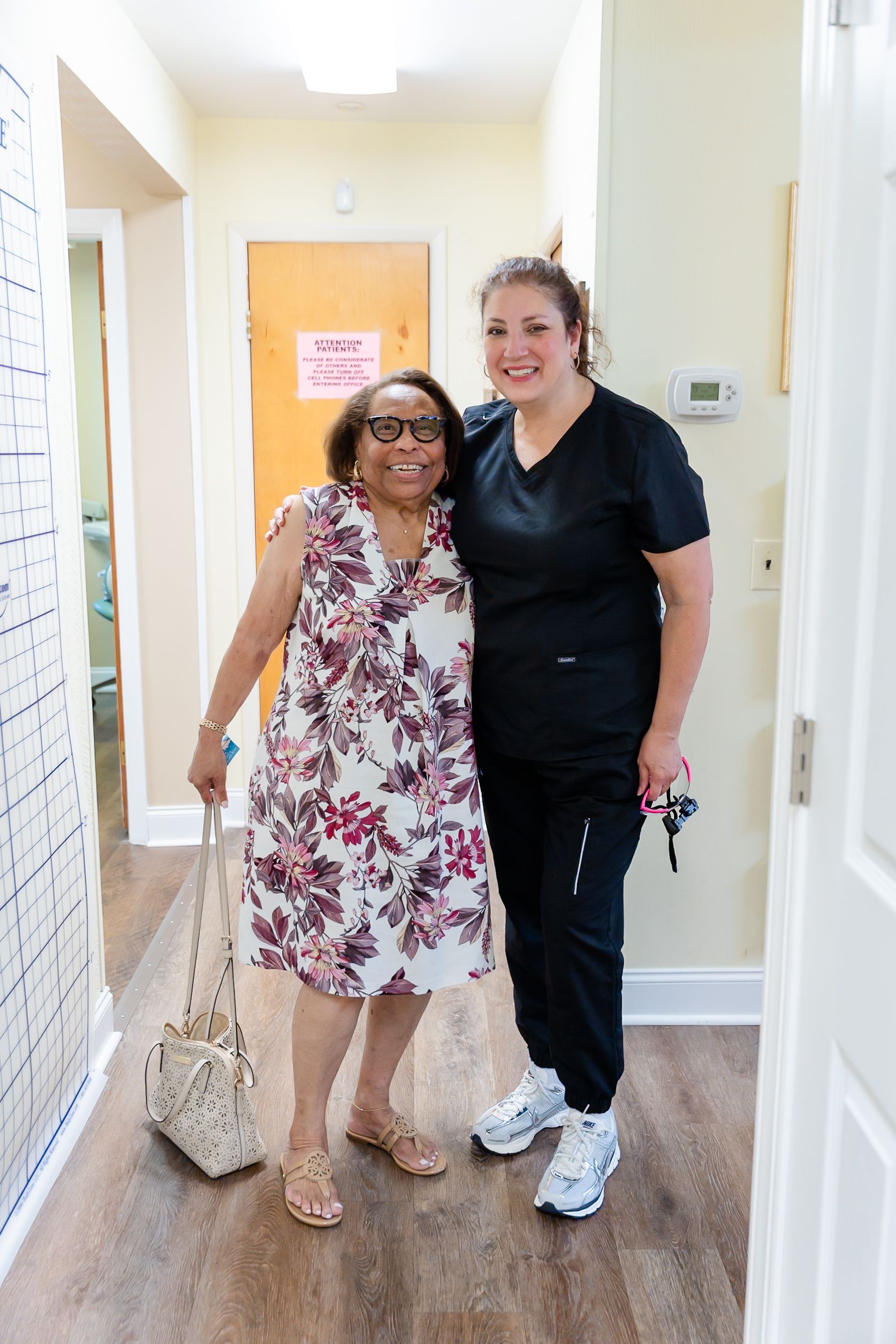 Woman in floral dress and woman in scrubs standing in hallway, smiling.