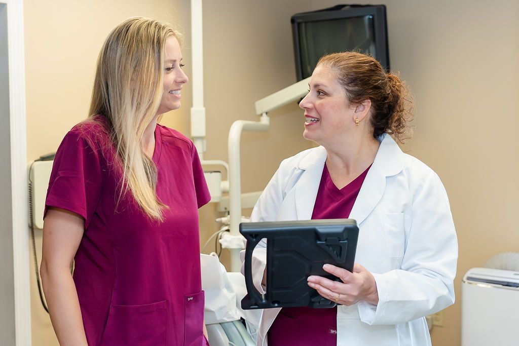 Two healthcare workers in a dental office; one in scrubs, one in a lab coat, are smiling and talking.