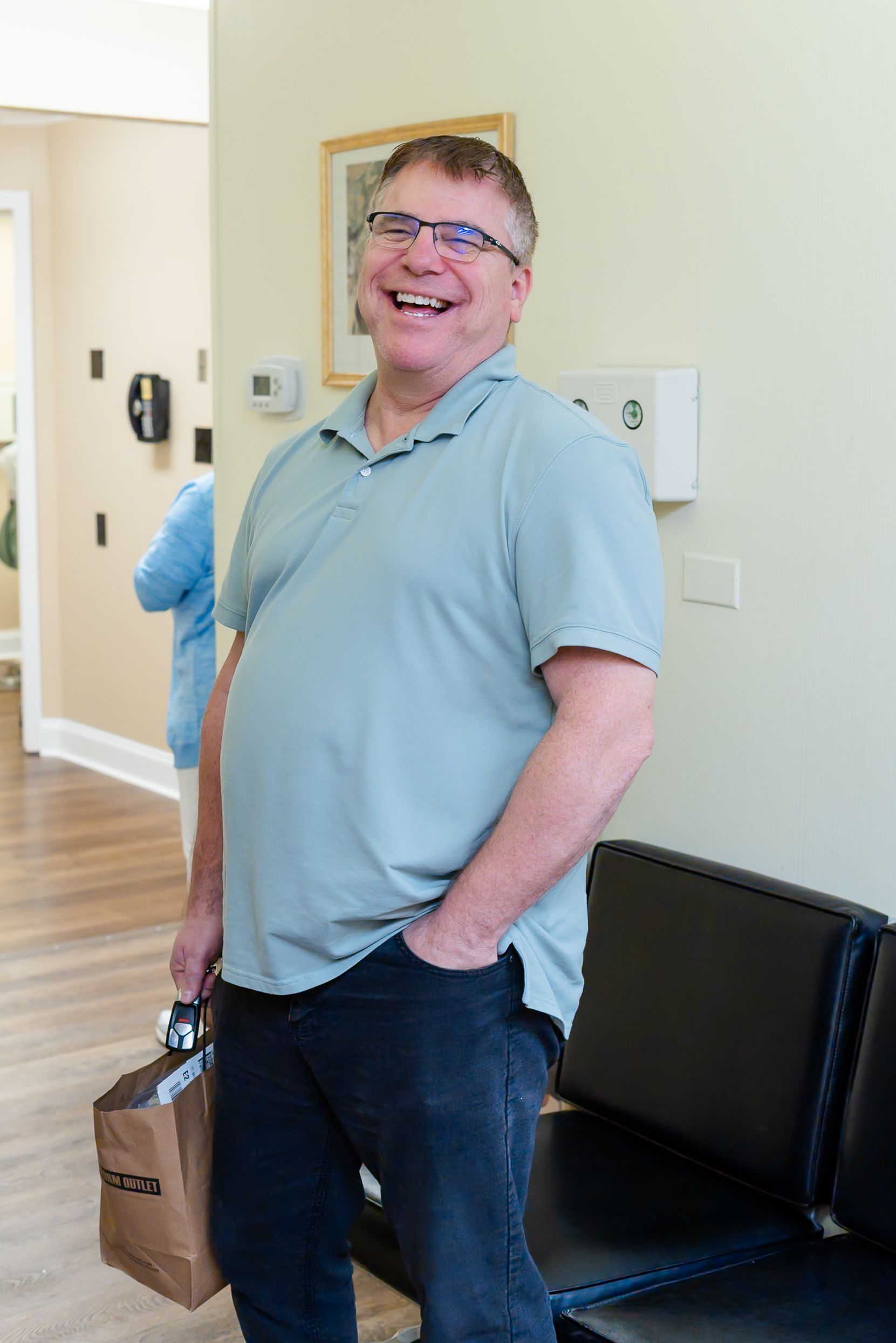 Man in a light green polo shirt, laughing, holding a bag, standing in an office.