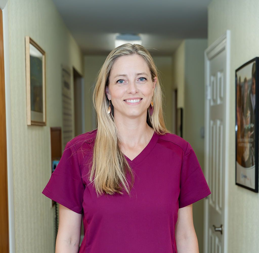 Woman in magenta scrubs smiles in a hallway, framed artwork on walls.