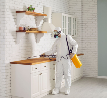 Person in white protective suit and mask spraying a kitchen countertop with a yellow sprayer.