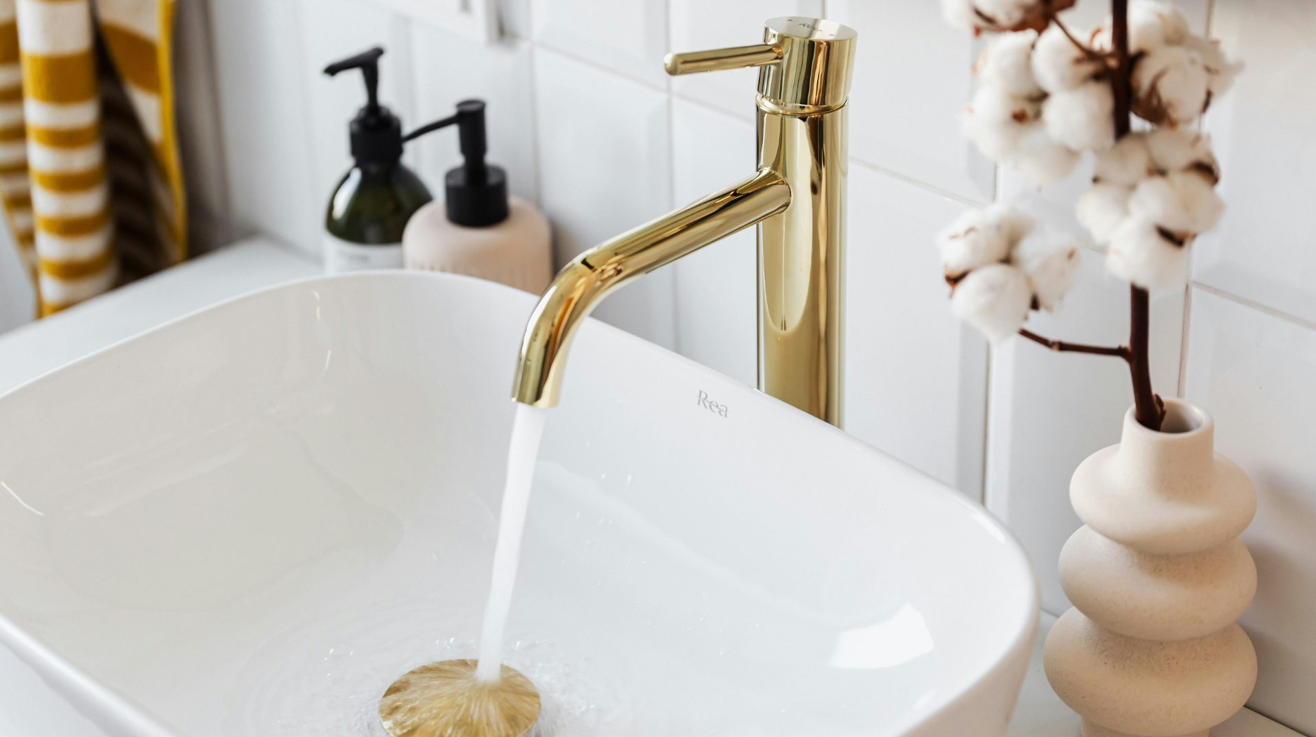 A bathroom sink with a gold faucet and a vase of cotton flowers.
