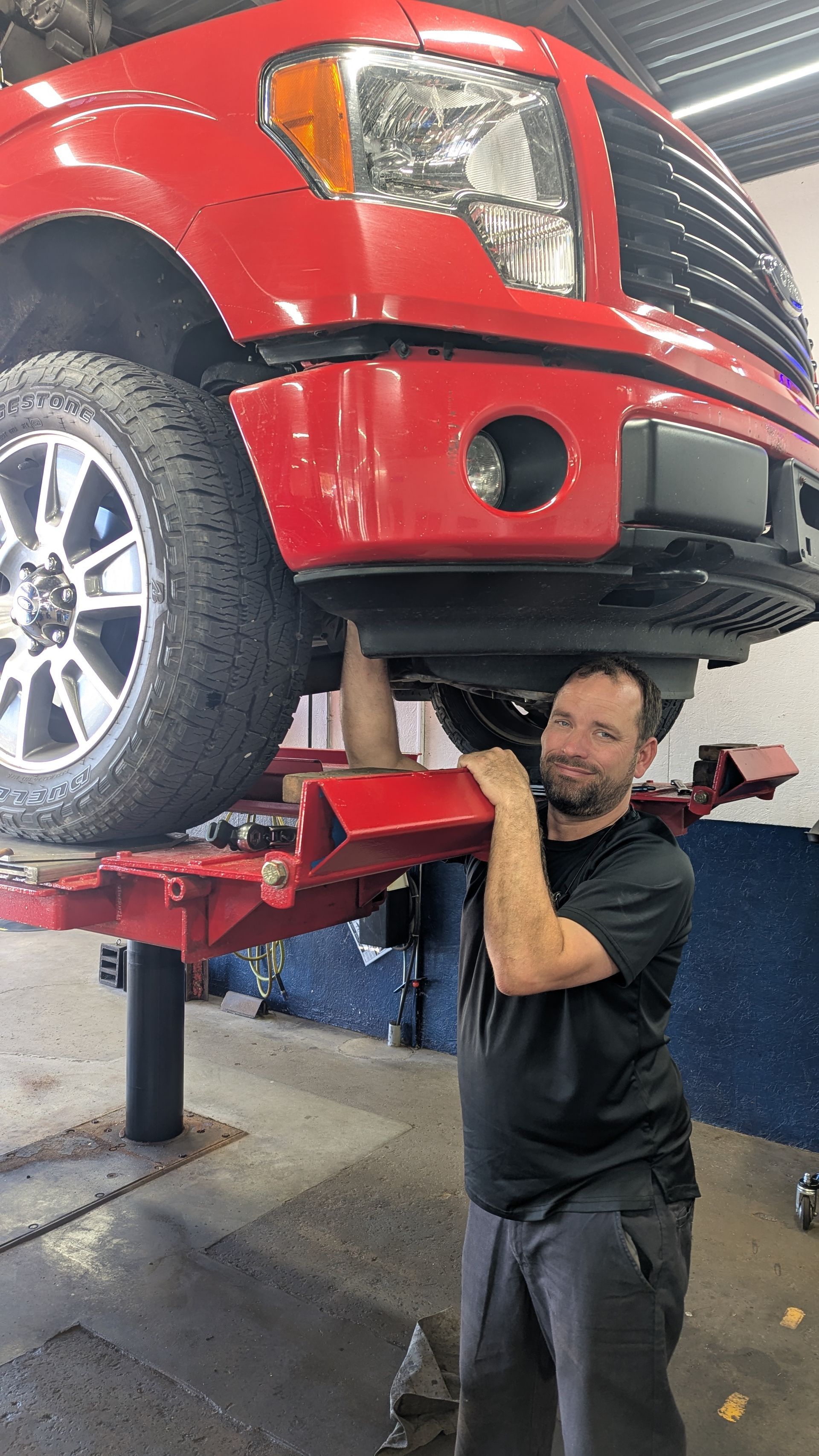 A mechanic stands in an auto shop, smiling while resting his arms on a red truck elevated on a hydraulic lift.