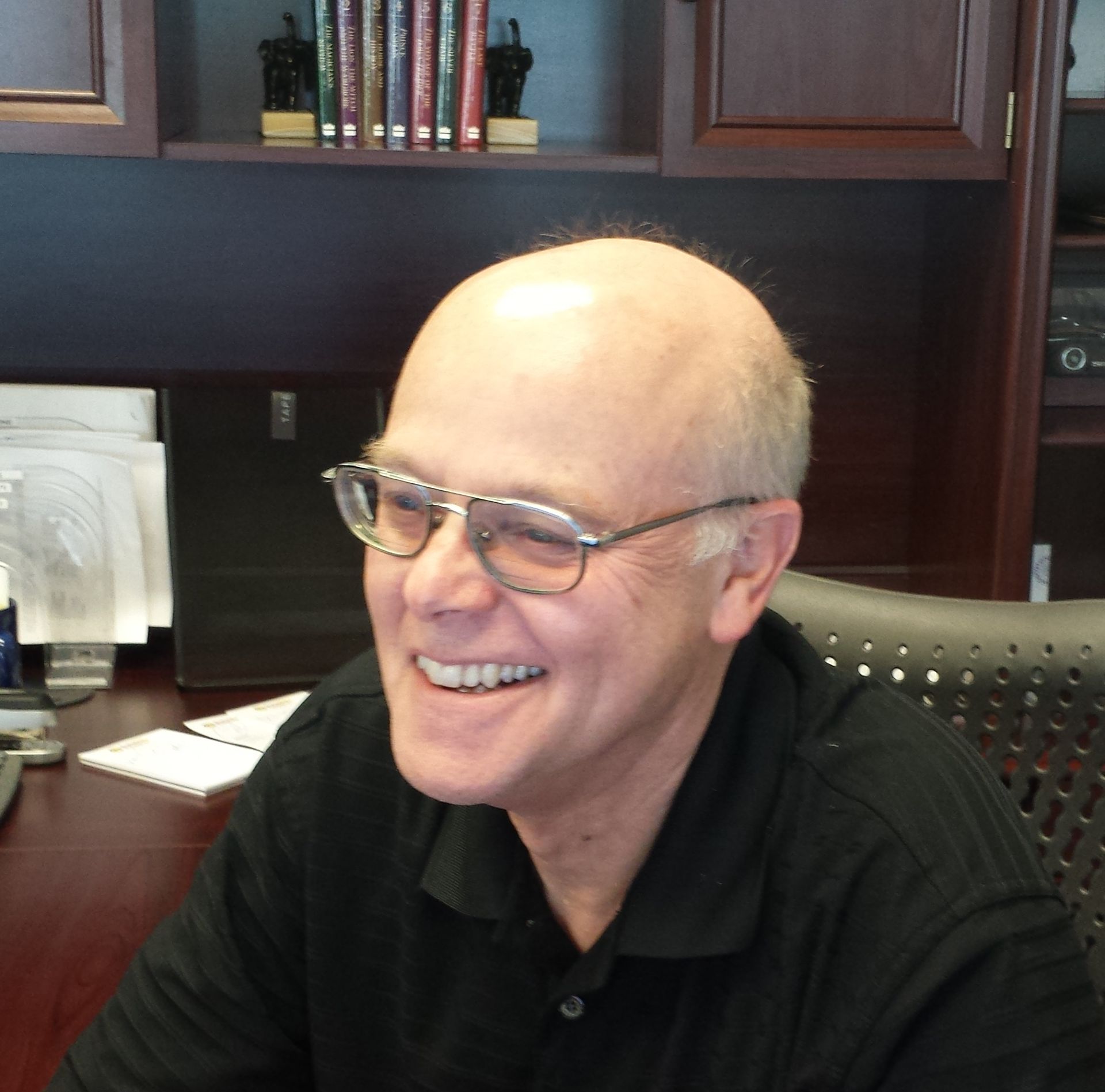 A smiling person wearing glasses and a black polo shirt, sitting at a desk in an office with bookshelves in the background.