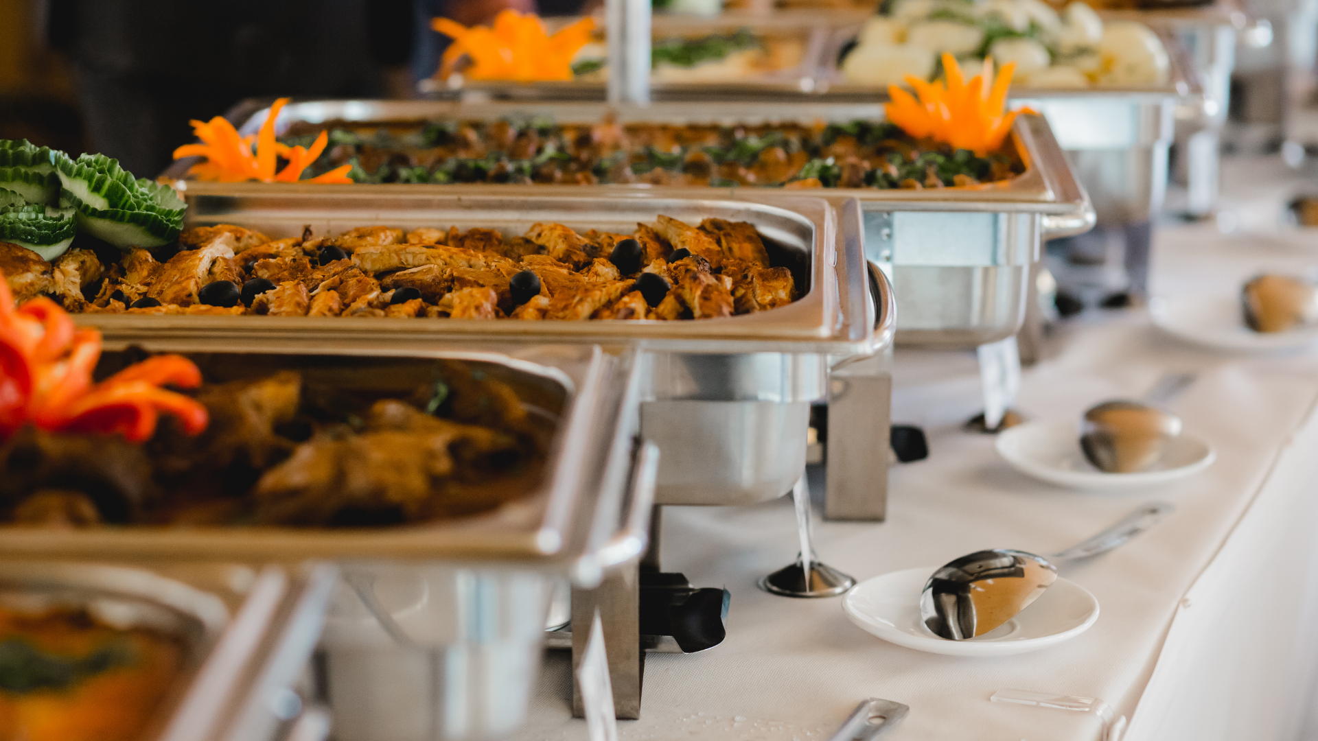 Buffet table with several hot dishes in stainless steel trays, decorated with orange flowers