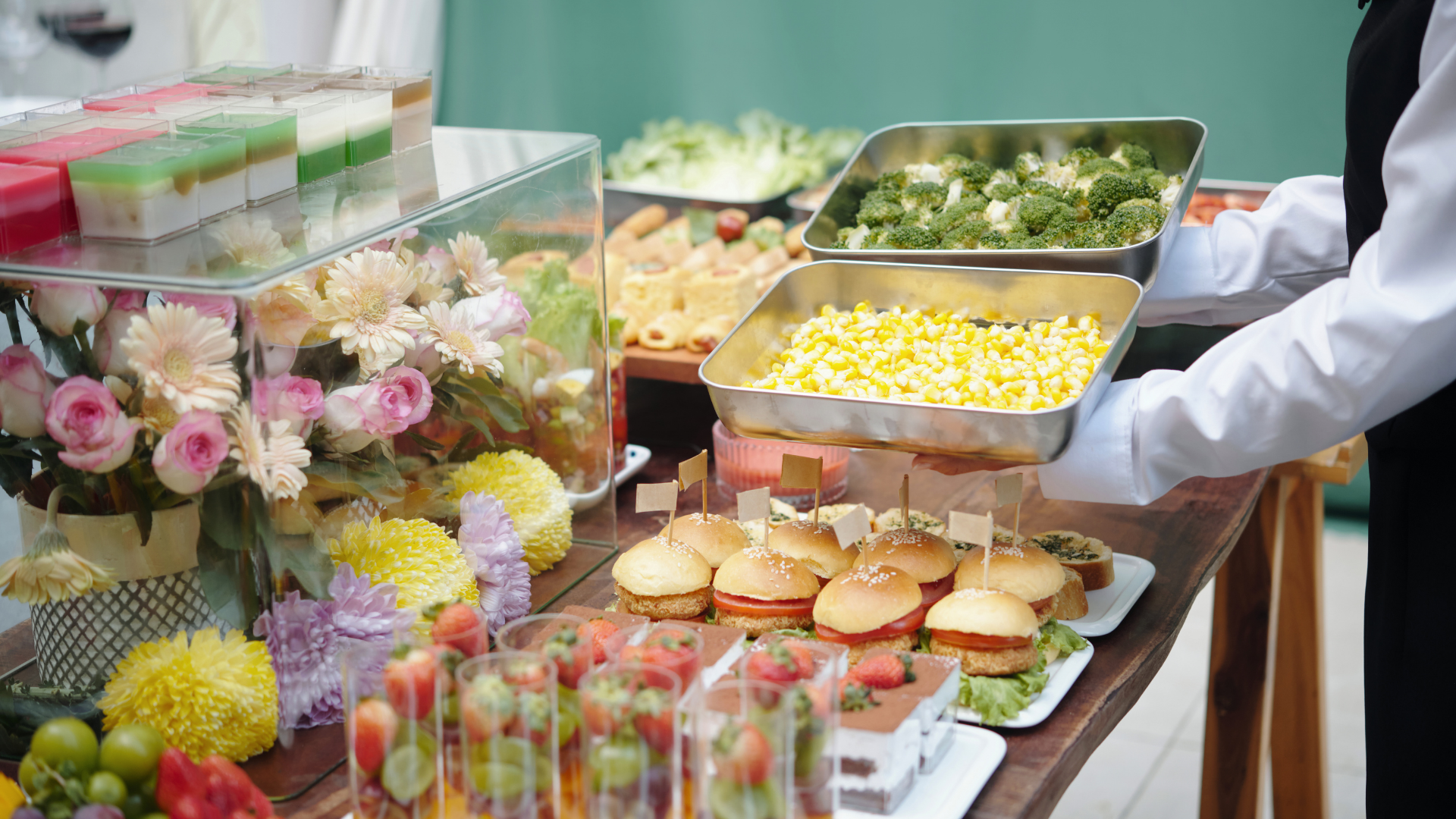 Buffet table with burgers, pastries, flowers, and trays of food at a catered event