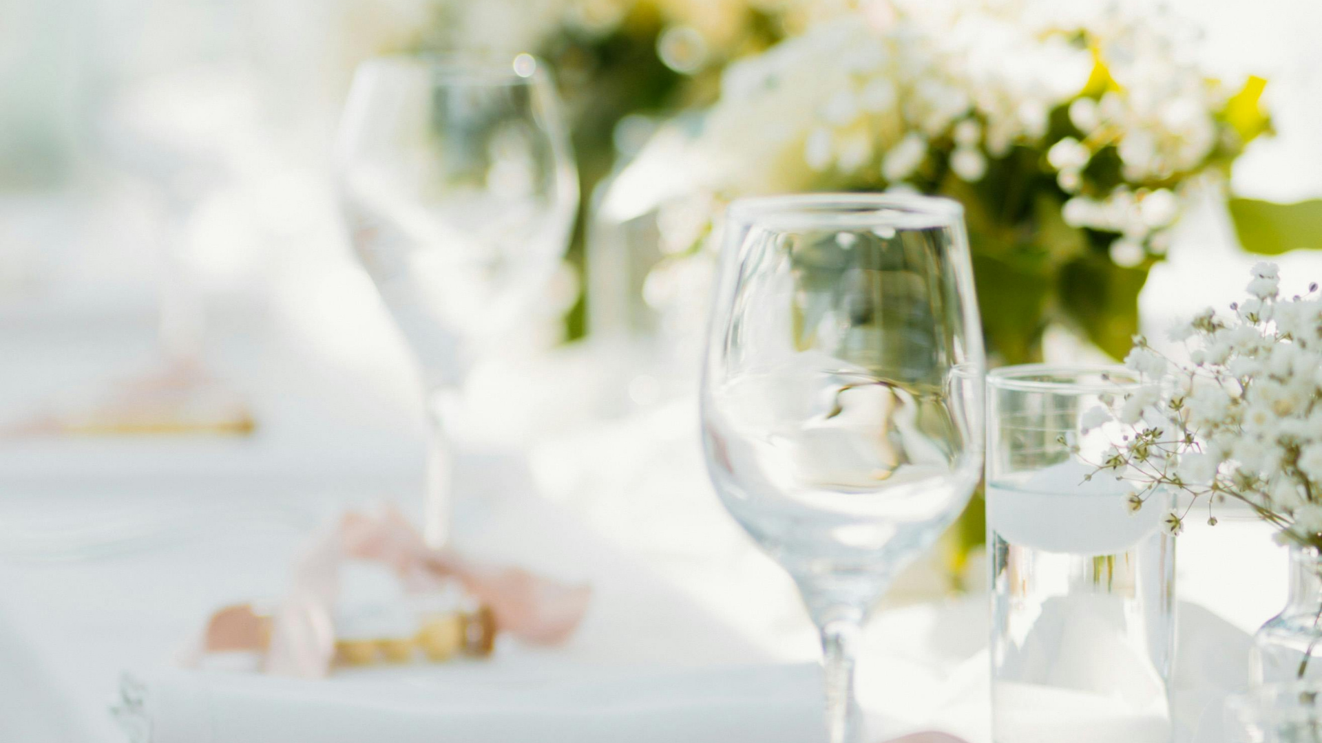 Elegant white wedding table setting with wine glasses, floral centerpieces, and soft sunlight