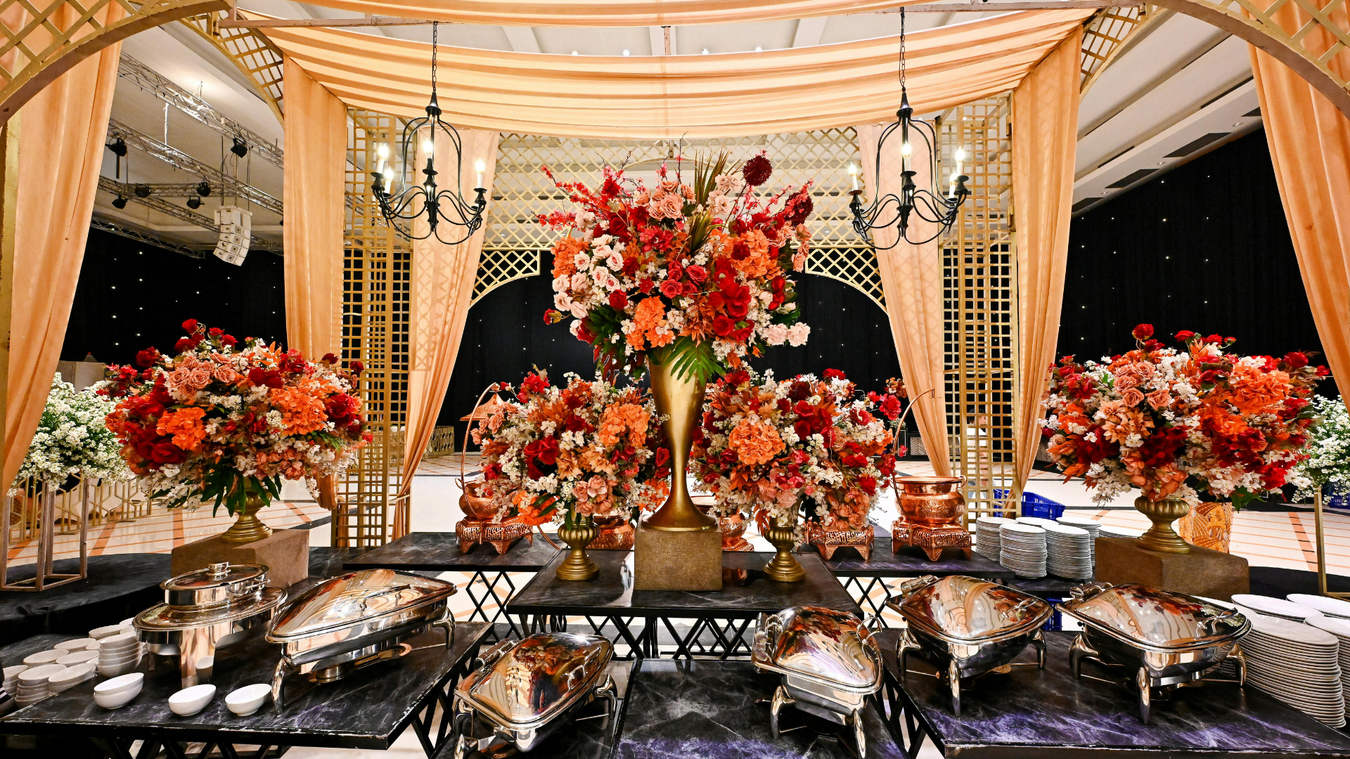 Elegant banquet table with silver serving dishes, red-orange floral centerpieces, and draped canopy lighting