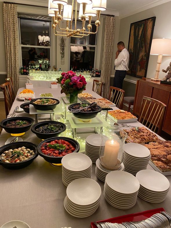 Elegant dining room with buffet spread, stacked plates, flower centerpiece, and a man standing by the wall.