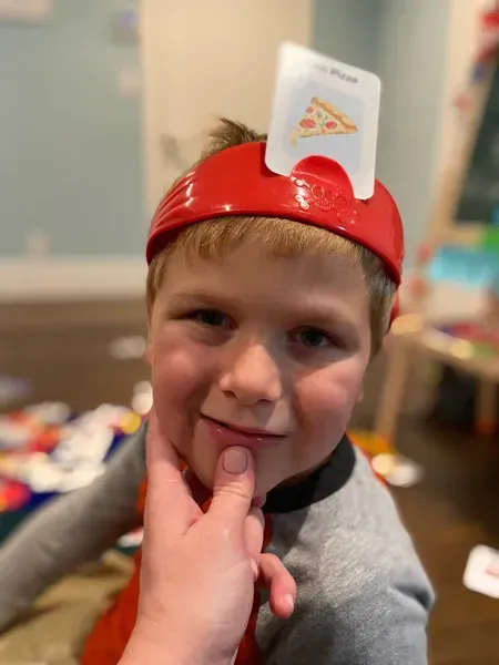 Child wearing a red hat with a card reading "Pizza" on it, smiling, with a finger touching his chin. Indoors.