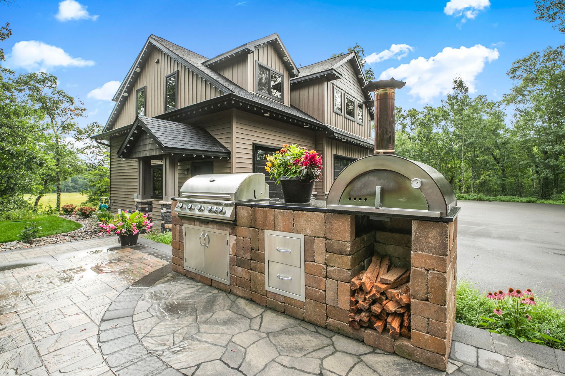 Stone outdoor pizza oven with a wood fire in front of a large house and driveway