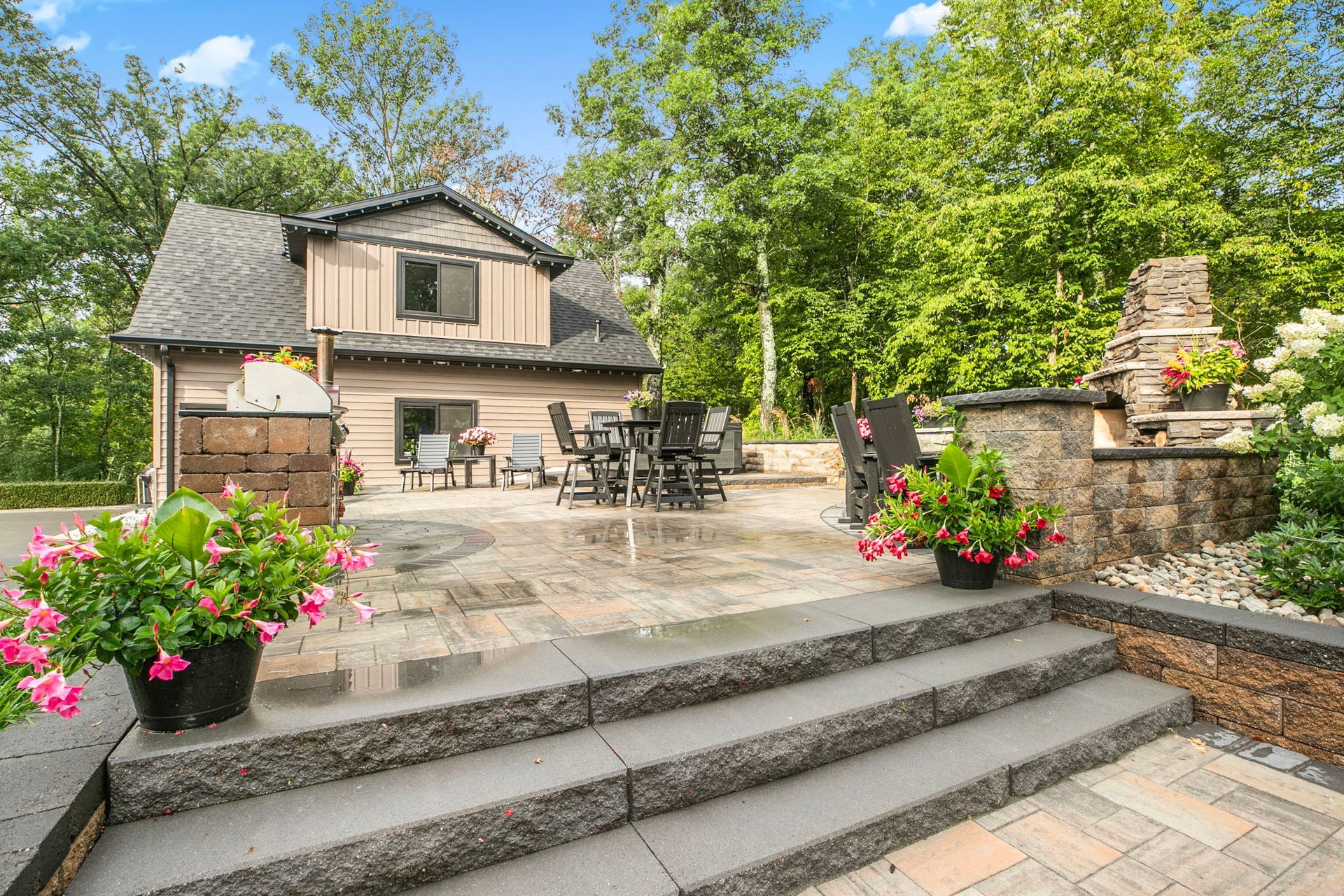 Stone patio with steps, outdoor seating, pink flowers, and a small house amid green trees