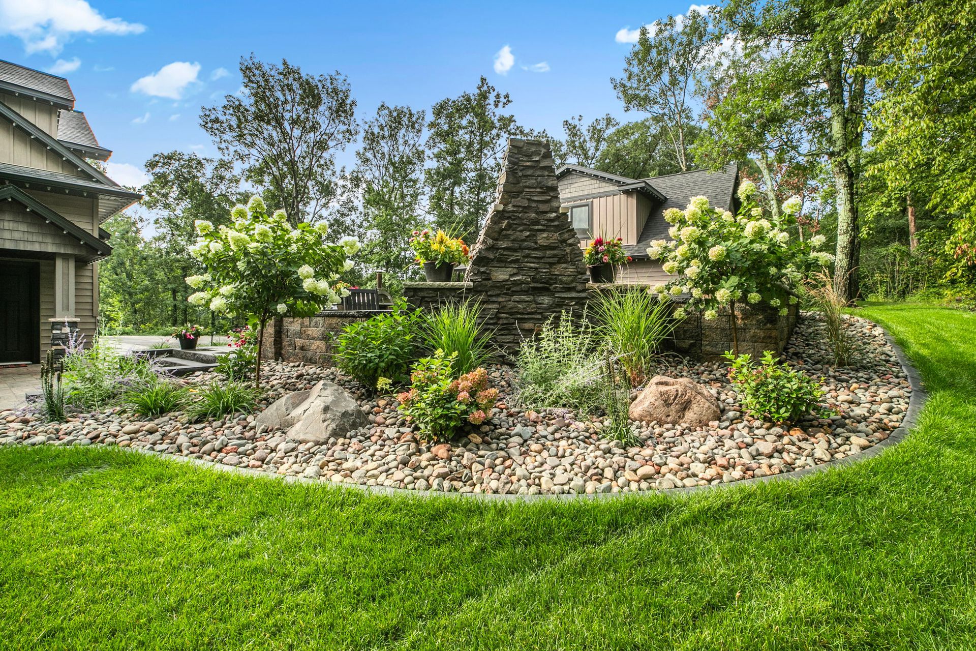 Landscaped garden with stone cairn, flowering shrubs, and green lawn beside a house