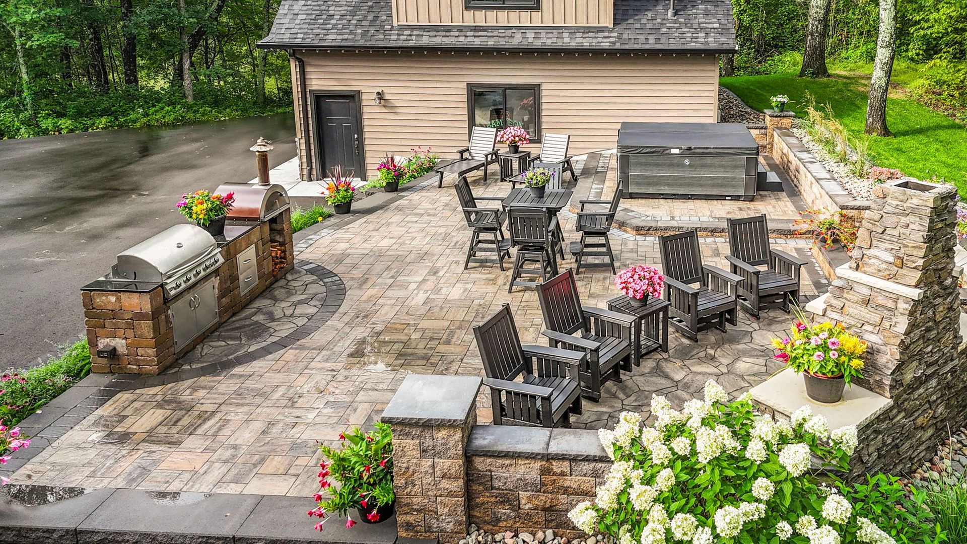 Stone patio with outdoor dining set, grill, and flower beds beside a house and stone wall