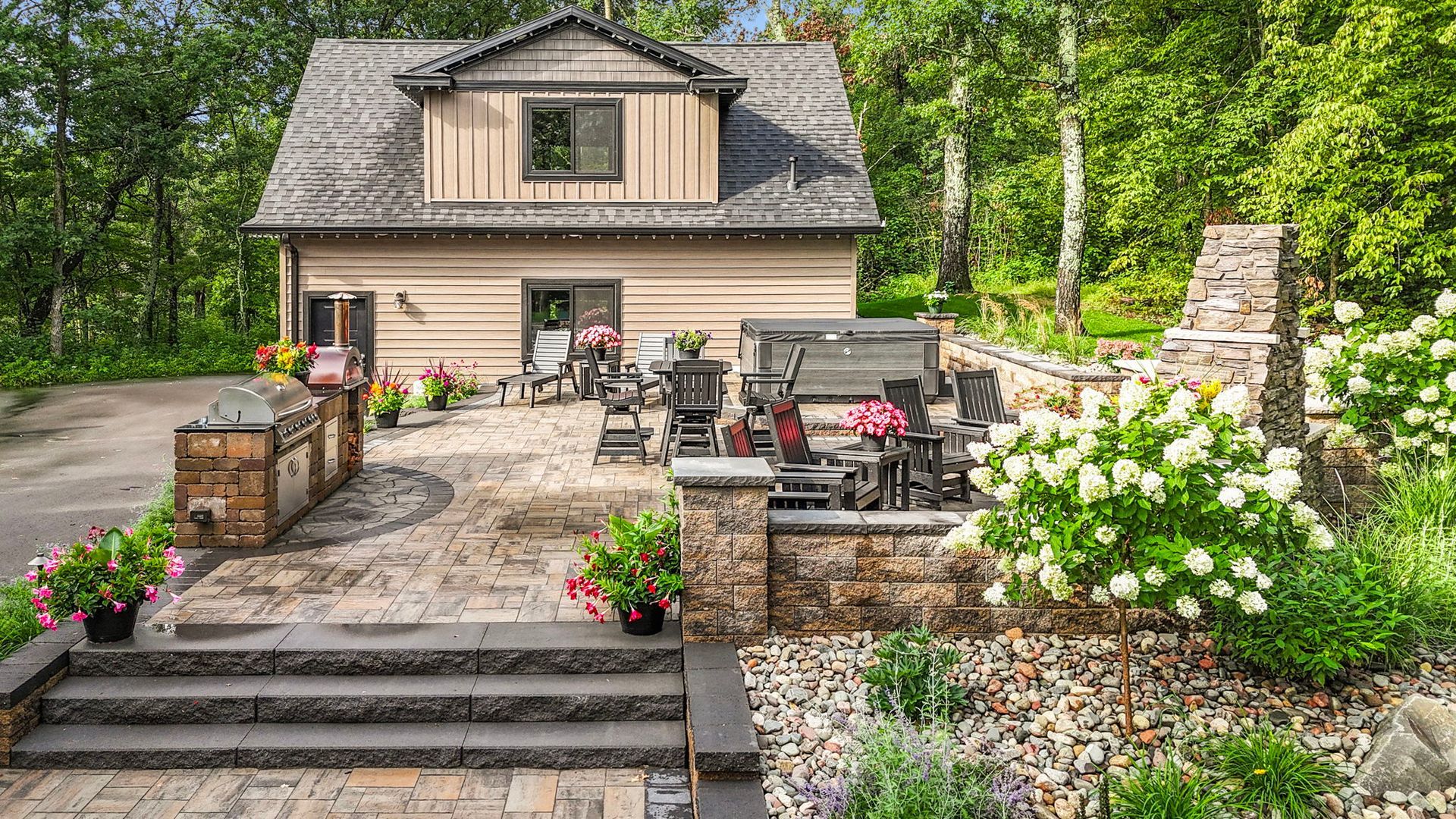 Backyard patio with stone steps, outdoor seating, flower beds, and a small cottage surrounded by trees.
